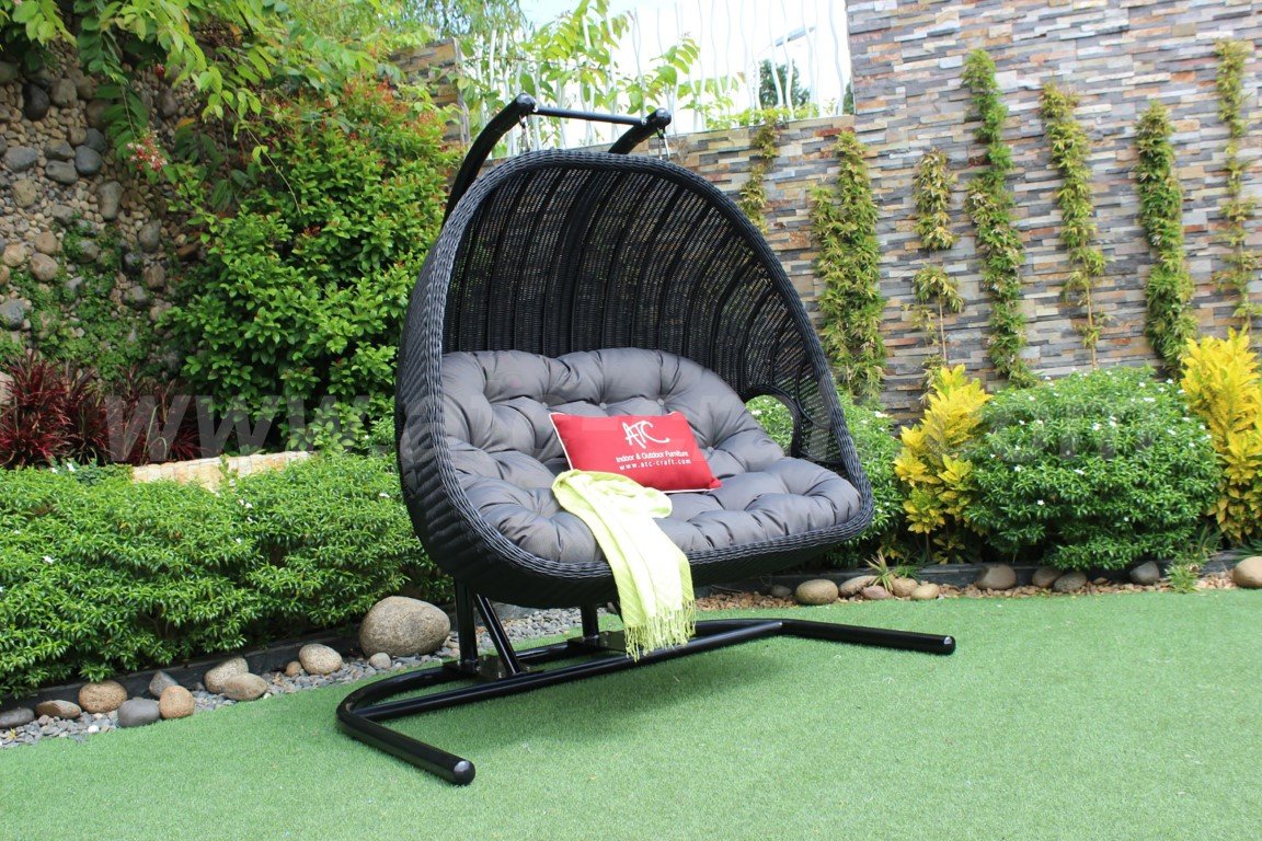 A modern synthetic rattan double swing chair with black wicker, gray cushions, and a red accent pillow, set on a green lawn with a backdrop of stone and greenery