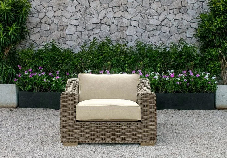 A stylish outdoor wicker armchair with beige cushions, placed on a gravel patio, set against a backdrop of greenery and a stone wall