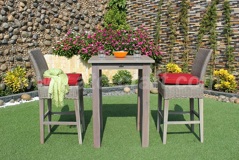 A stylish outdoor bar set featuring a tall wooden table and two wicker bar chairs with red cushions, placed on artificial grass in a garden with a decorative stone wall and vibrant flowers