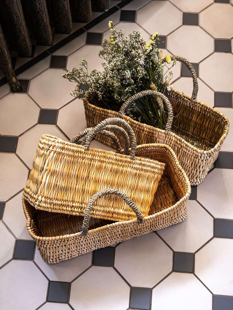 Two woven rattan baskets with handles placed on a black and white tiled floor. One basket contains a bouquet of delicate flowers, adding a natural touch to the decor